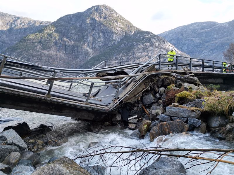 Ei bru over ei elv med steiner under. Brua har kollapsa og har ein knekk på midten. Bak brua er det høge fjell.