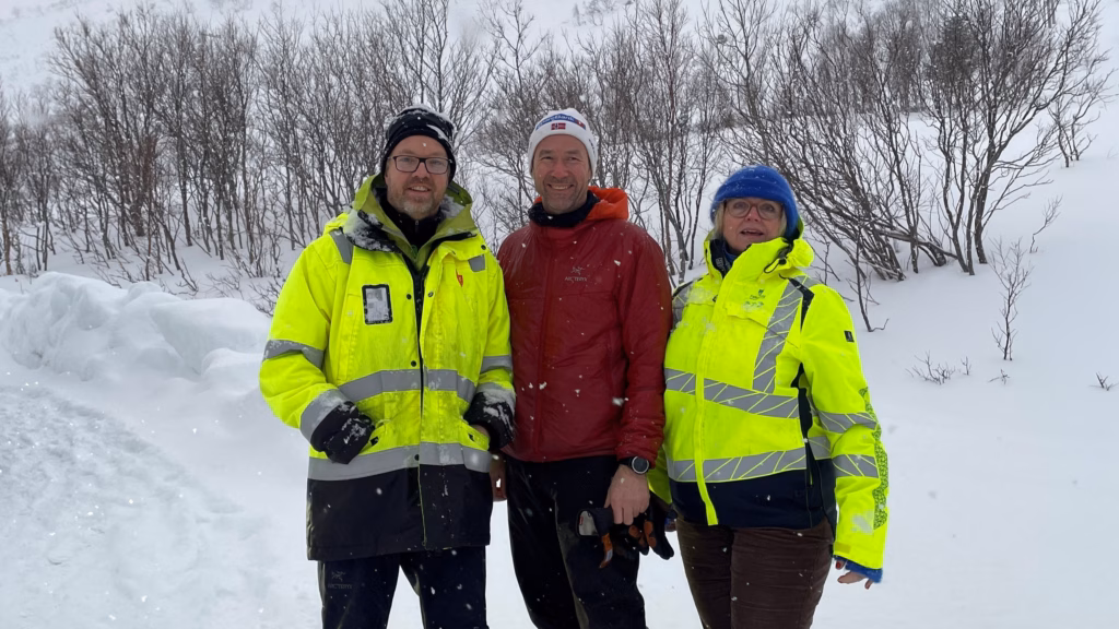 Tre personer smiler mot kamera. Mannen til venstre har på gul jakke og mørk lue, mannen i midten har hvit lue og rød jakke, kvinnen til høyre har gul jakke og blå lue på. Bak dem er det trær og snø. 