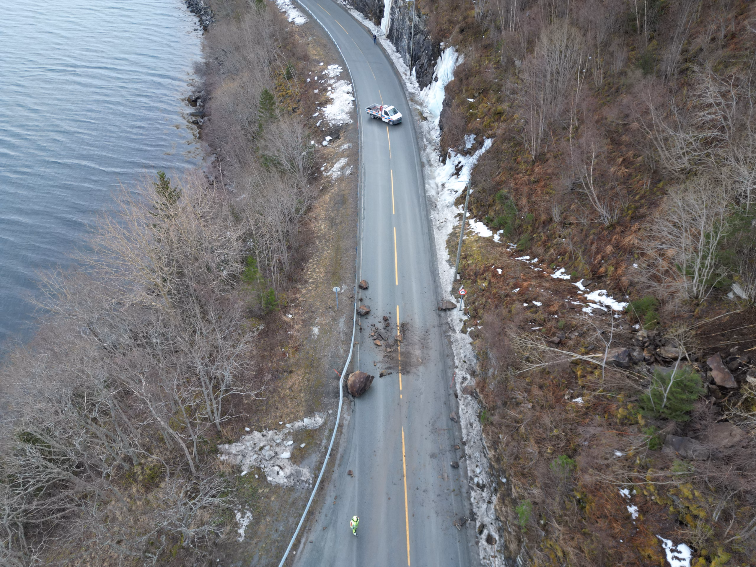 En grå vei med fjell på ene siden og sjø på andre siden. På veien ligger flere steiner. En bil står parkert et stykke unna raset.