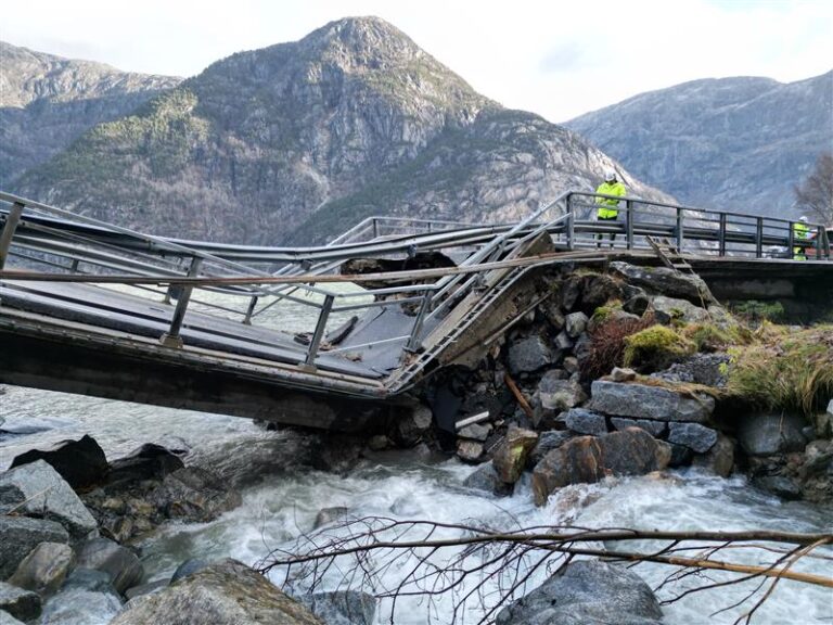 Ei bru over ei elv med steiner under. Brua har kollapsa og har ein knekk på midten. Bak brua er det høge fjell.